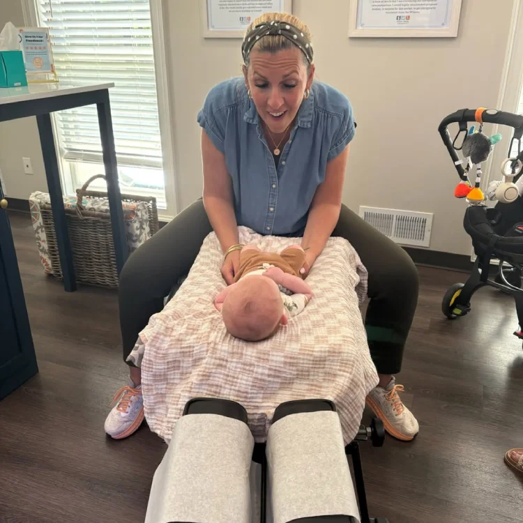 Female chiropractor smiles while holding infant on treatment table