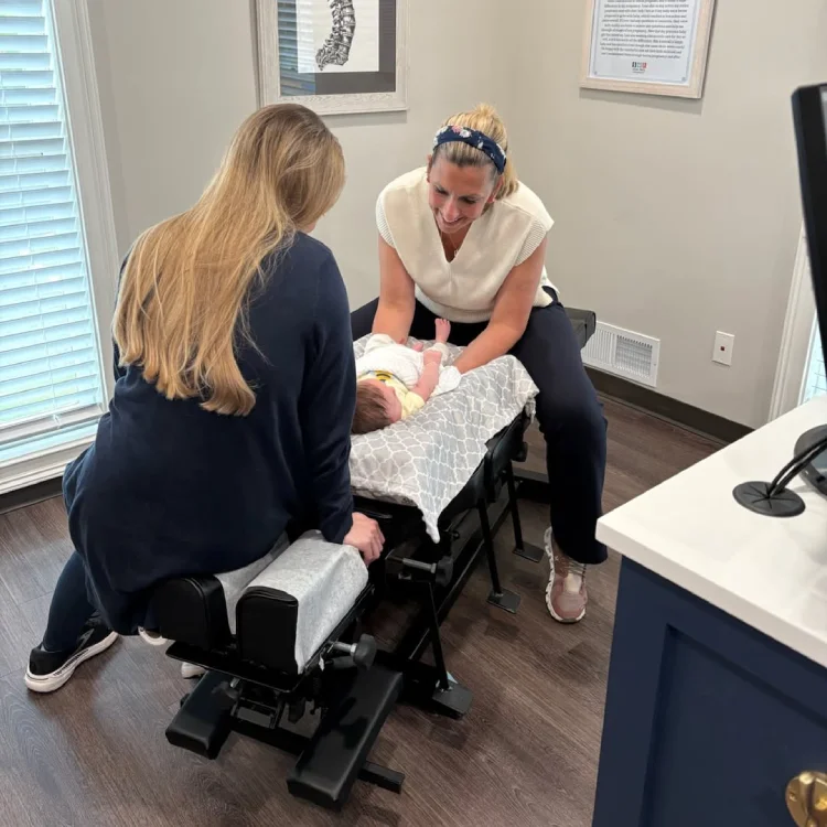 Female chiropractor assesses infant on treatment table while smiling and mother observes from other side of treatment table
