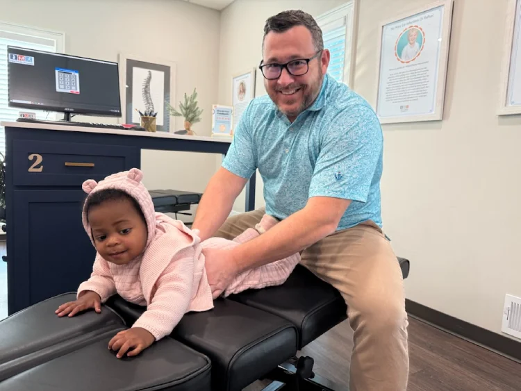 Male chiropractor smiles while holding infant on treatment table