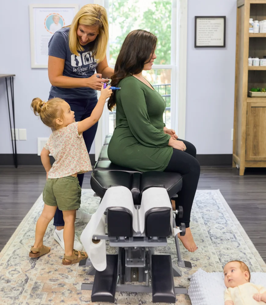 Woman sits on treatment table while her young daughter and the chiropractor playfully assess her back